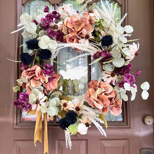 Puede incluir: Una corona floral decorativa con flores melocotón, moradas y crema, acentuada con follaje verde y una cinta dorada. La corona se exhibe en una puerta marrón.
