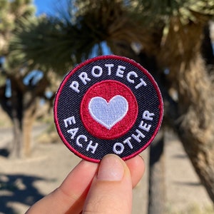 May include: A black and red round embroidered patch with a white heart in the center. The text on the patch reads "Protect Each Other".