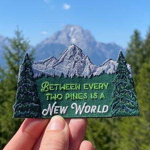 May include: Green and white embroidered patch with the text "Between every two pines is a new world" against a backdrop of a mountain range and a lake.