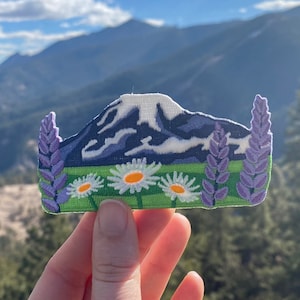 May include: An embroidered patch featuring a mountain range with purple lupine flowers and white daisies. The patch is held in front of a mountainous backdrop.