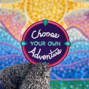 Może przedstawiać: Haftowany naszywka w kolorze fioletowym i różowym z wzorem kompasu. Tekst na naszywce brzmi "Choose Your Own Adventure".