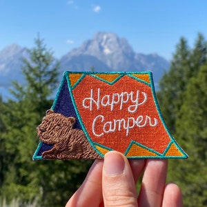 Könnte beinhalten: Ein gestickter Aufnäher mit einem braunen Bären, der aus einem Zelt schaut. Das Zelt ist orange und gelb mit dem Text "Happy Camper" in weiß.