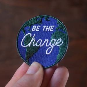 Puede incluir: Parche bordado redondo con un diseño de globo azul y continentes verdes. El texto blanco dice "BE THE CHANGE". El parche está sujeto por una mano, con un fondo marrón.