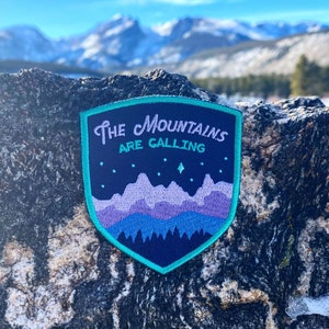 May include: A blue and purple embroidered patch with the text "The Mountains Are Calling" and a stylized mountain range with stars. The patch is on a dark gray rock.