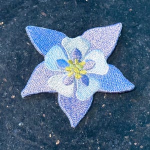 Könnte beinhalten: Ein gestickter Blumen-Patch in Blau und Weiß mit gelber Mitte. Die Blume hat fünf Blütenblätter und ist ein beliebtes Design für Kleidung und Accessoires.
