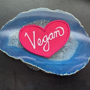 Può includere: Un cerotto rosa a forma di cuore con la parola "Vegan" scritta in lettere bianche corsive. Il cerotto è appoggiato su una pietra di agata blu e bianca.