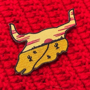 Peut inclure: Un patch brodé représentant un crâne jaune avec un coucher de soleil et des cactus en arrière-plan.