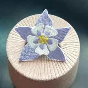 May include: A purple and white embroidered flower with yellow center, resembling a columbine, is displayed on a light brown textured surface.