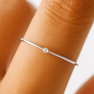 May include: A delicate silver ring with a small, clear gemstone at the center. The ring is thin and minimalist, showcasing a simple yet elegant design. The gemstone sparkles, adding a touch of subtle brilliance to the jewelry.
