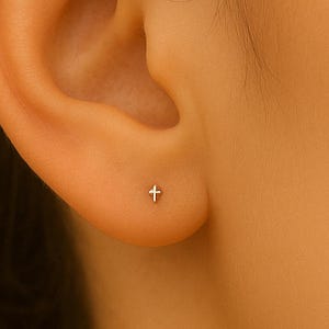 May include: A close-up of a person's ear with a small, silver cross stud earring. The earring is centered on the earlobe. The background is a warm, neutral tone.