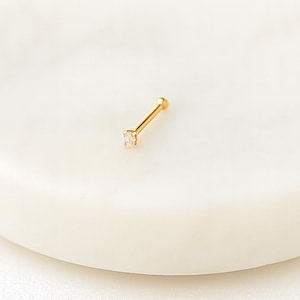 May include: A gold-colored nose stud with a small, square, clear gemstone. The stud is set on a white, circular surface. The jewelry is simple and elegant.