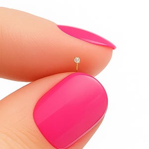 May include: A tiny, gold-colored nose stud with a single, clear, round gemstone. The stud is held between two fingers with bright pink nail polish against a white background. The jewelry is delicate and minimalist.