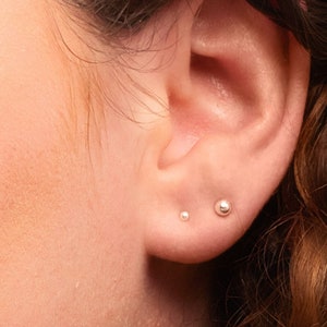 May include: Close-up of a person's ear with two small silver stud earrings. The earrings are round and have a smooth finish.