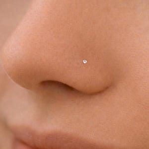 May include: Close-up of a person's face with a small, sparkling nose stud. The stud is a tiny, round, clear gem set in a metal base. The skin is smooth and fair, with a subtle glow. The focus is on the nose and the delicate jewelry.