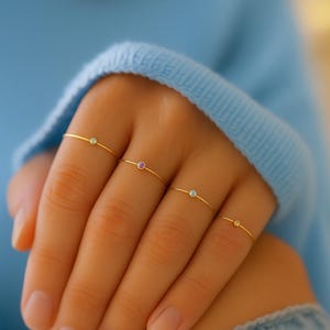 May include: Four delicate gold rings, each featuring a small, colorful gemstone. The gemstones are a light blue, pink, and clear. The rings are displayed on a hand, with a light blue sweater in the background.