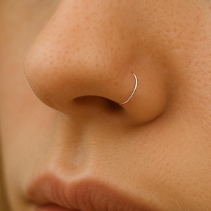 May include: Close-up of a person's face with a silver nose ring. The ring is a thin, curved piece of metal, and it sits in the nostril. The skin is fair with a slight pink hue. The person's lips are slightly parted.
