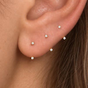 May include: Five small, round, clear stud earrings are arranged in a curved line along the earlobe and lower cartilage. Each earring features a single, tiny, brilliant-cut stone set in a simple, polished gold-tone metal setting, creating a delicate look.