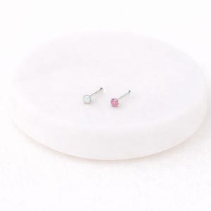 May include: Two nose studs with small, square gemstones. One stud features a light blue gemstone, and the other has a pink gemstone. Both are set on thin, silver-colored posts. The studs are displayed on a white, circular surface.