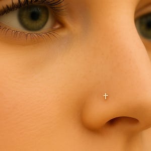 May include: Close-up of a person's face with a small, silver cross nose stud. The skin tone is light, and the background is a warm, neutral color. The focus is on the nose and the delicate jewelry.