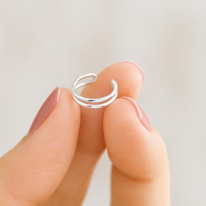 May include: A silver-colored ear cuff with a double-band design. The cuff is held between fingers, showcasing its minimalist and modern style. The jewelry piece is designed to be worn on the ear without piercing.