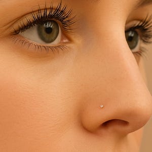 May include: Close-up of a person's face, focusing on the nose with a small, round nose stud. The skin is a light tone, and the eyes are green with long, dark eyelashes. The image is well-lit, highlighting the details of the face.