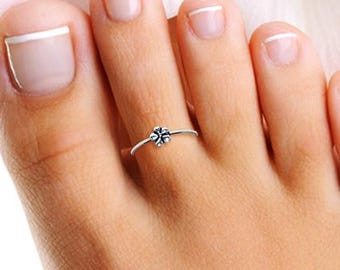 May include: A silver toe ring featuring a small, flower-shaped design. The ring is delicate and sits on a person's toe. The background is a soft, blurred white.