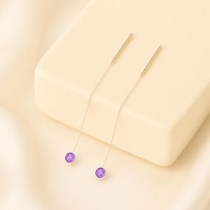 May include: A pair of silver threader earrings with a small, round, purple gemstone at the end of each chain. The earrings are displayed on a cream-colored surface, with a soft, neutral background.
