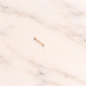 May include: A rose gold industrial barbell piercing. The straight barbell has a small ball on each end. The jewellery is displayed on a white and grey marble surface.