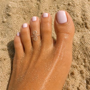 May include: A silver-colored metal toe ring featuring a double spiral design. The ring consists of a thin wire that curves into two opposing circular coils, creating an open-ended band that rests on the second toe of a foot with light pink nail polish.