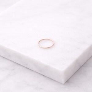 K&ouml;nnte beinhalten: Ein zarter ros&eacute;goldener Ring, platziert auf einer wei&szlig;en Marmoroberfl&auml;che. Der Ring ist ein schlichtes Band, das ein minimalistisches Design zeigt. Der warme Farbton des Rings kontrastiert mit dem k&uuml;hlen wei&szlig;en Hintergrund und unterstreicht seine elegante Schlichtheit.