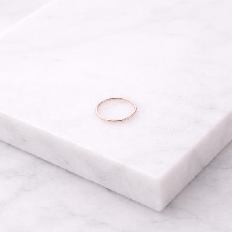 K&ouml;nnte beinhalten: Ein zarter ros&eacute;goldener Ring, platziert auf einer wei&szlig;en Marmoroberfl&auml;che. Der Ring ist ein schlichtes Band, das ein minimalistisches Design zeigt. Der warme Farbton des Rings kontrastiert mit dem k&uuml;hlen wei&szlig;en Hintergrund und unterstreicht seine elegante Schlichtheit.