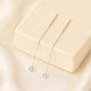 May include: A pair of silver threader earrings. Each earring features a delicate chain with a small, round, light blue gemstone at the end. The earrings are displayed on a cream-colored surface, creating a minimalist and elegant aesthetic.