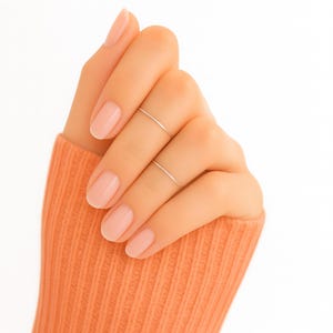 May include: A hand wearing two thin, silver rings. The nails are painted a light pink. The person is wearing an orange sweater. The background is white.