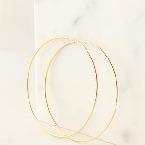 Peut inclure: Une paire de grandes créoles dorées. Les boucles d'oreilles sont fines et circulaires, avec une finition lisse et polie. Elles sont posées sur un fond de marbre blanc, créant une esthétique minimaliste.