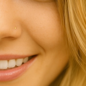 May include: Close-up of a smiling person's face, with a small, round, gold nose stud. The skin is a warm, light tone, and the hair is blonde. The focus is on the nose and mouth.