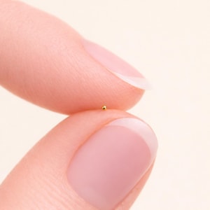 May include: A tiny, gold-colored bead held between two fingers. The bead is perfectly round and reflects light, appearing shiny against the skin. The image is a close-up, highlighting the small size and detail of the bead.