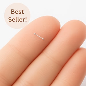 May include: A small, silver nose stud with a straight bar and two small, round ends. The text "Best Seller!" is in a light brown circle in the background.