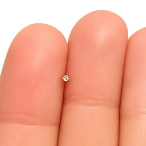 May include: A tiny gold nose stud with a small, clear gemstone. The stud is held between two fingers against a white background. The jewelry is delicate and minimalist.