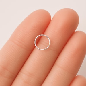 May include: A small, silver-colored nose ring, held between two fingers. The ring is a simple, thin circle with a small gap. The skin tone is light, and the background is a soft, neutral color.