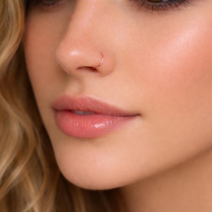 K&ouml;nnte beinhalten: Nahaufnahme eines Gesichts, das einen kleinen Nasenring aus Ros&eacute;gold zeigt. Der Ring ist ein einfacher, d&uuml;nner Reifen. Die Haut hat einen nat&uuml;rlichen Glanz, mit zarten rosa Lippen und einem Hauch von Rouge.