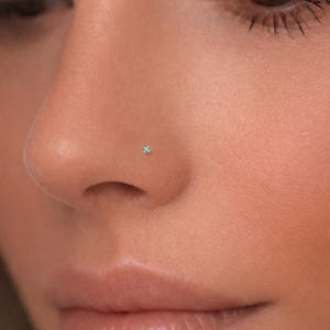 May include: Close-up of a nose with a small, square, light blue nose stud. The skin tone is fair, and the focus is on the nose piercing. The image is well-lit, highlighting the jewelry.