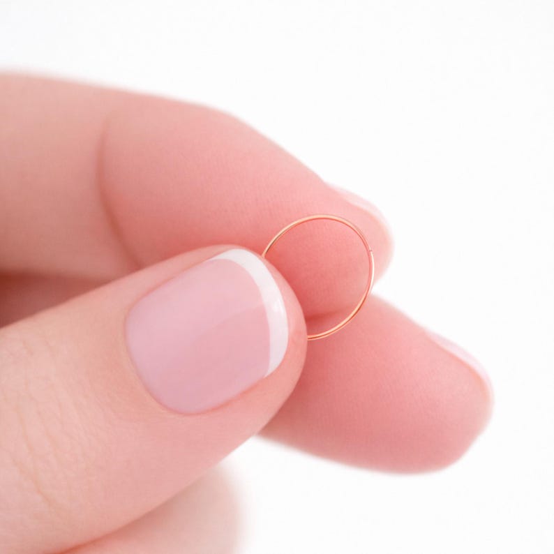 K&ouml;nnte beinhalten: Ein kleiner, goldfarbener Creolen-Ohrring. Der Ohrring wird zwischen zwei Fingern vor einem wei&szlig;en Hintergrund gehalten. Der Ohrring hat ein einfaches, kreisf&ouml;rmiges Design und scheint aus d&uuml;nnem Metall zu bestehen.