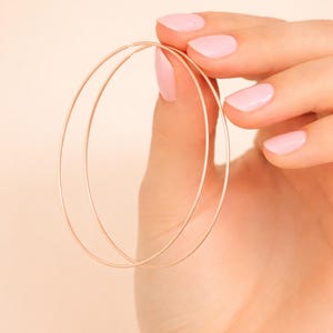 May include: A pair of large, thin, gold-colored hoop earrings. The earrings are held in a hand with pink nail polish against a soft, peach-colored background. The hoops are simple and elegant.