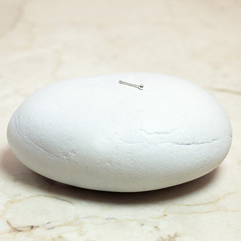 May include: A small silver nose stud with a round ball on the end. The stud is sitting on a white rock.