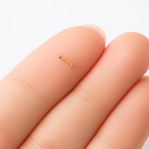 May include: A small, gold-colored nose bone piercing. The jewelry features a straight bar with a small ball on each end. The piercing is shown on a finger, against a white background.