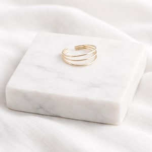 May include: A gold-colored ring with three parallel bands. The open-ended design allows for adjustable sizing. The ring is displayed on a white marble block, set against a white fabric background.