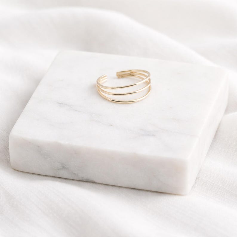 May include: A gold-colored ring with three parallel bands. The open-ended design allows for adjustable sizing. The ring is displayed on a white marble block, set against a white fabric background.