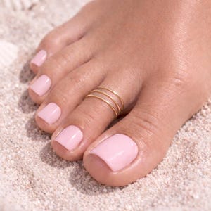 May include: A close-up of a foot with a gold toe ring. The ring has three thin bands. The toenails are painted a light pink color. The foot is resting on a sandy surface.