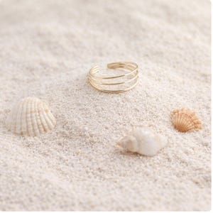 May include: A gold-colored ring with three bands rests on white sand, near seashells. The ring has a simple, elegant design. The scene evokes a beach or coastal theme.