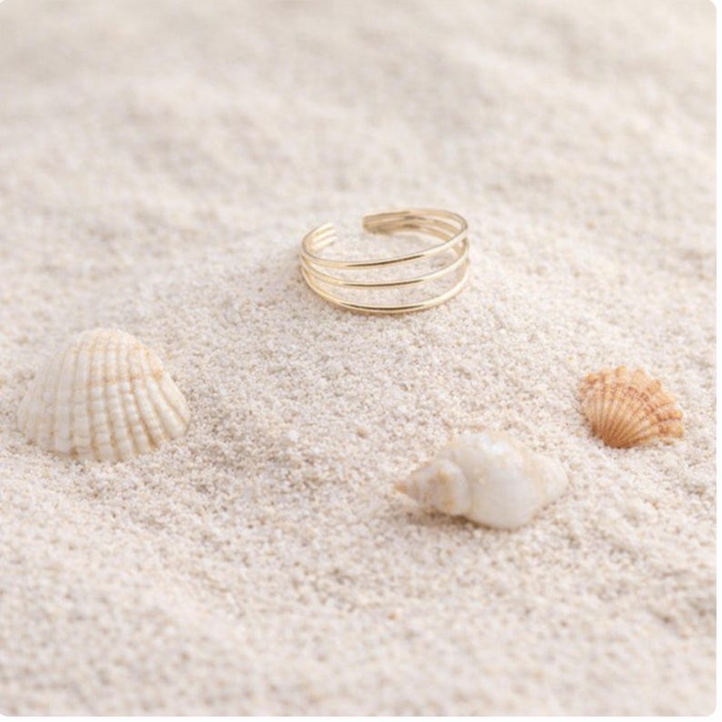 May include: A gold-colored ring with three bands rests on white sand, near seashells. The ring has a simple, elegant design. The scene evokes a beach or coastal theme.